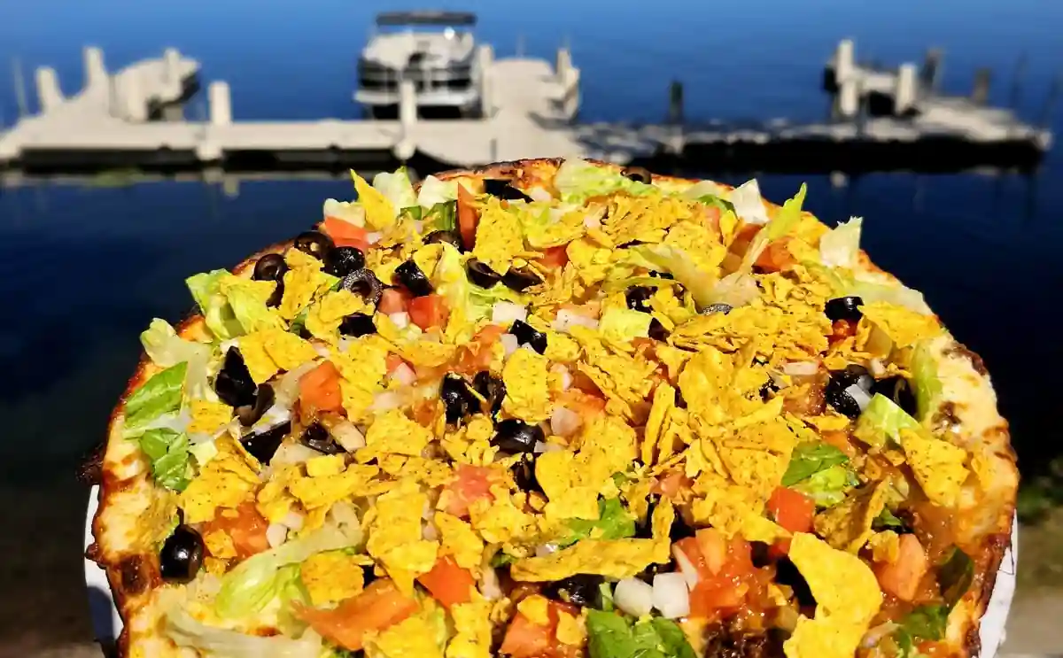 A Pioneer taco pizza held up over the dock with a pontoon and Long Lake behind